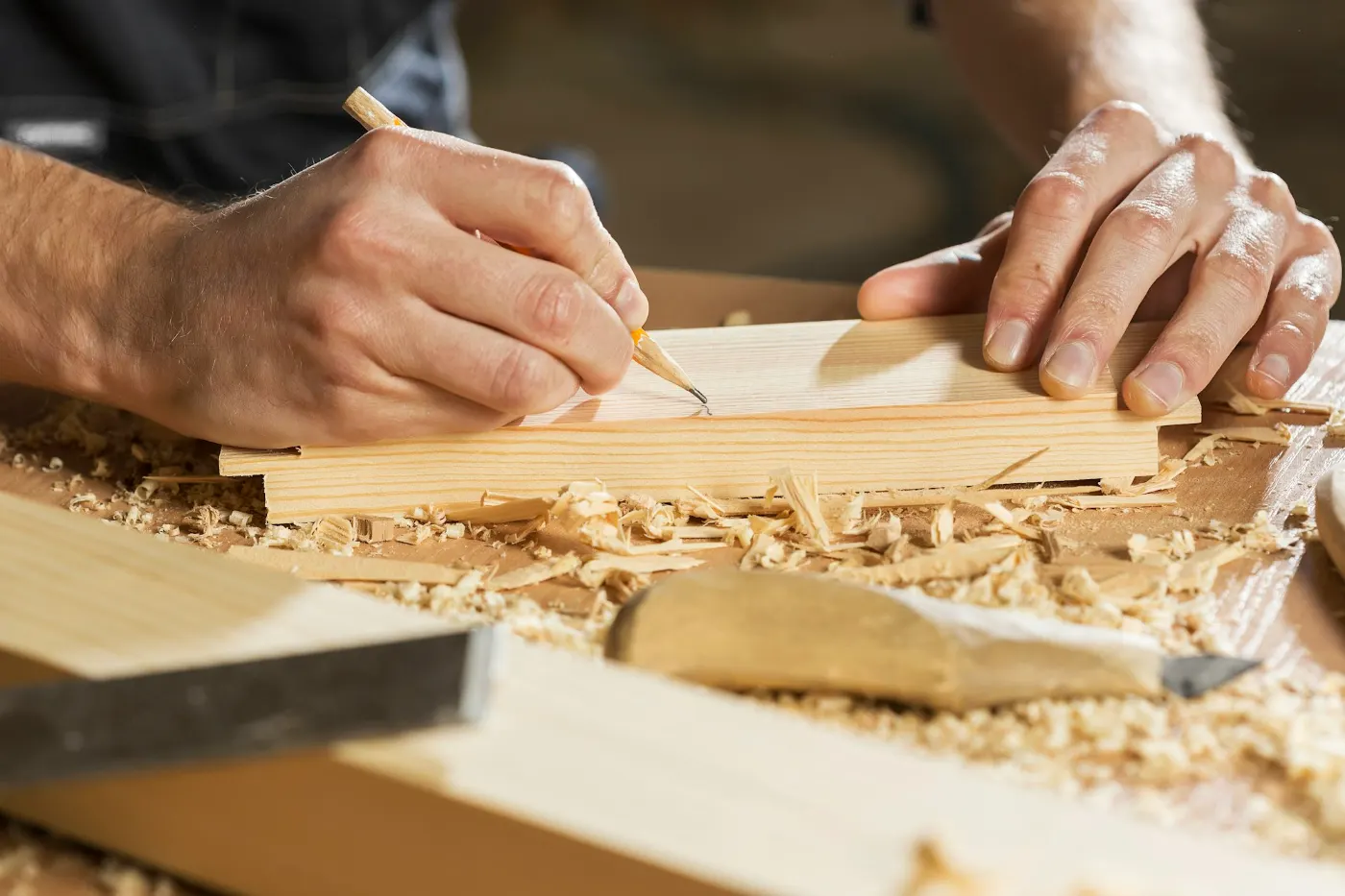 Hände reißen mit dem Bleistift eine Linie auf einem Holzbrett an, daneben Hobelspäne.